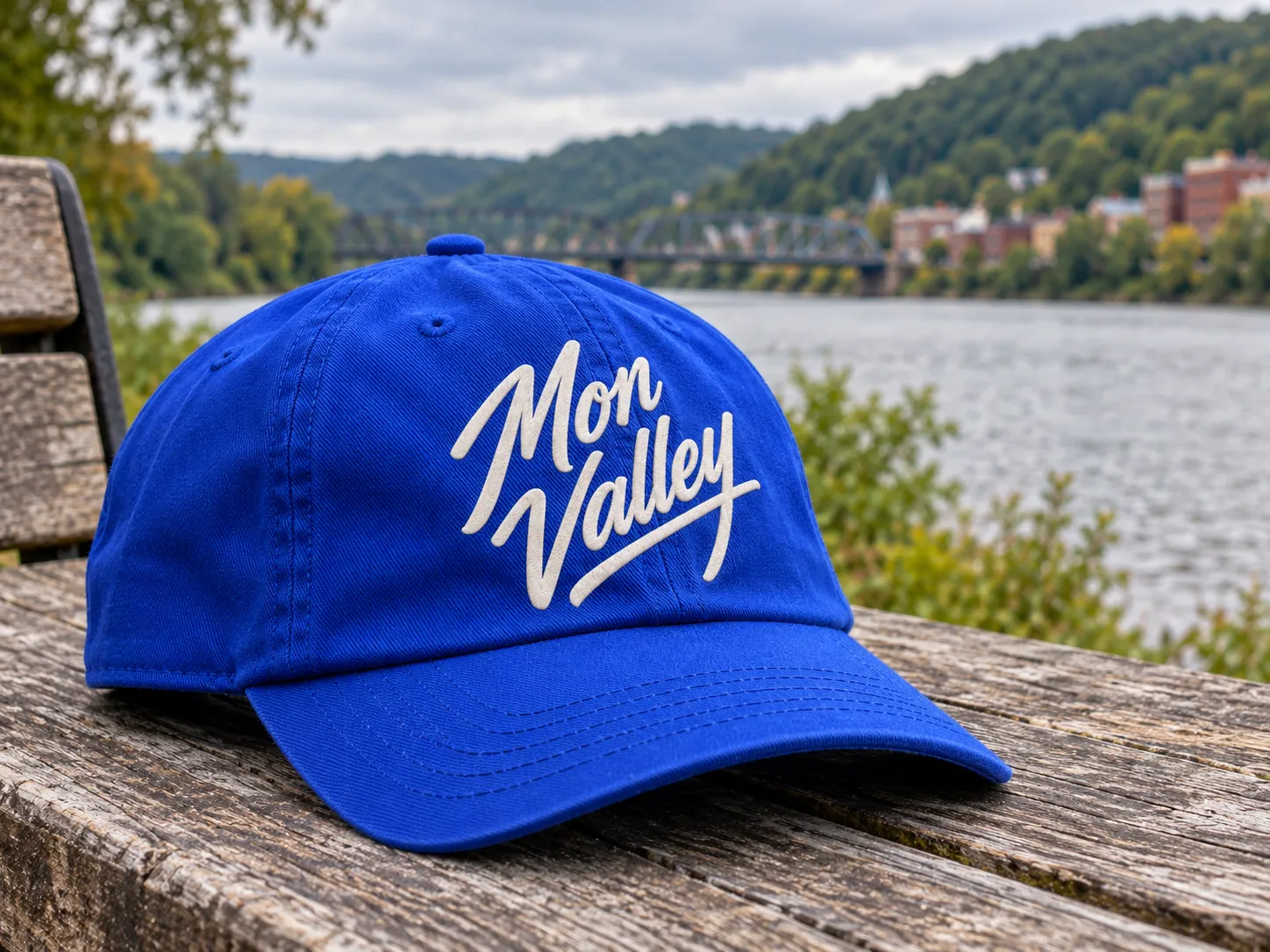 A cobalt blue cap with the Mon Valley logo embroidered in cream, on a bench by the river